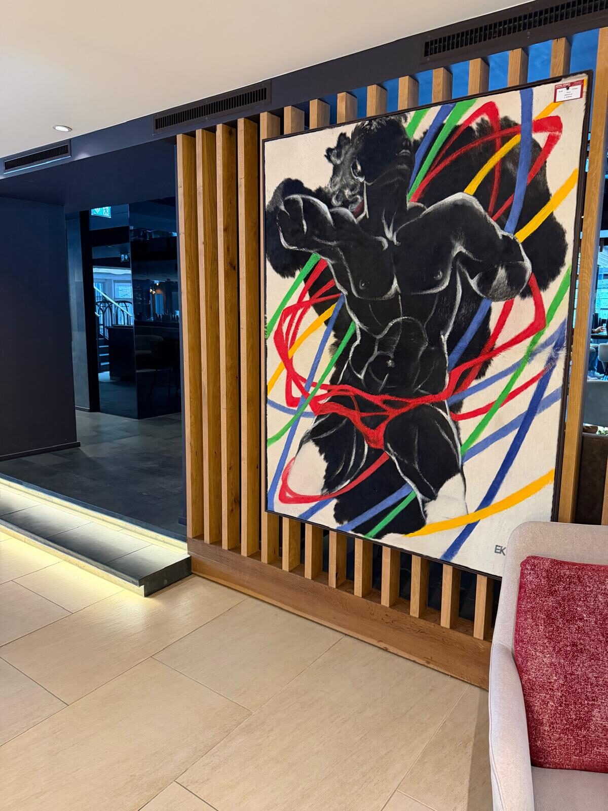 Großformatiges Fellbild einer dynamischen schwarzen Figur mit farbigen Bändern, installiert an einer Holzlamellen-Wand im Lebenberg Schlosshotel in Kitzbühel