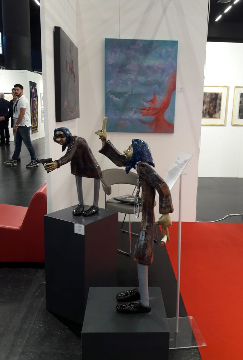 Drahtskulpturen auf Sockeln vor Gemäldewand am Messestand