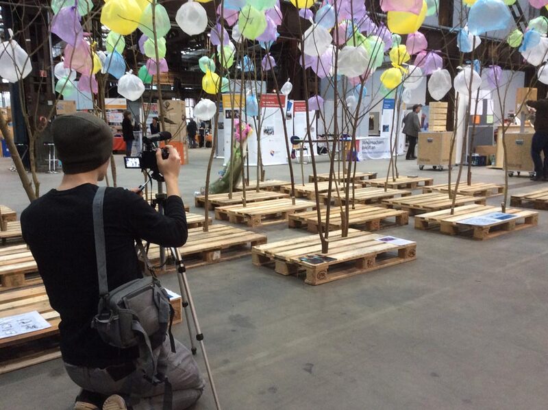Bäumchen auf Holzpaletten mit bunten Beuteln in einer Messehalle