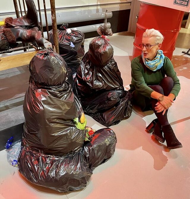 Die Künstlerin sitzt neben zwei Figuren aus schwarzen Müllsäcken im Ausstellungsraum