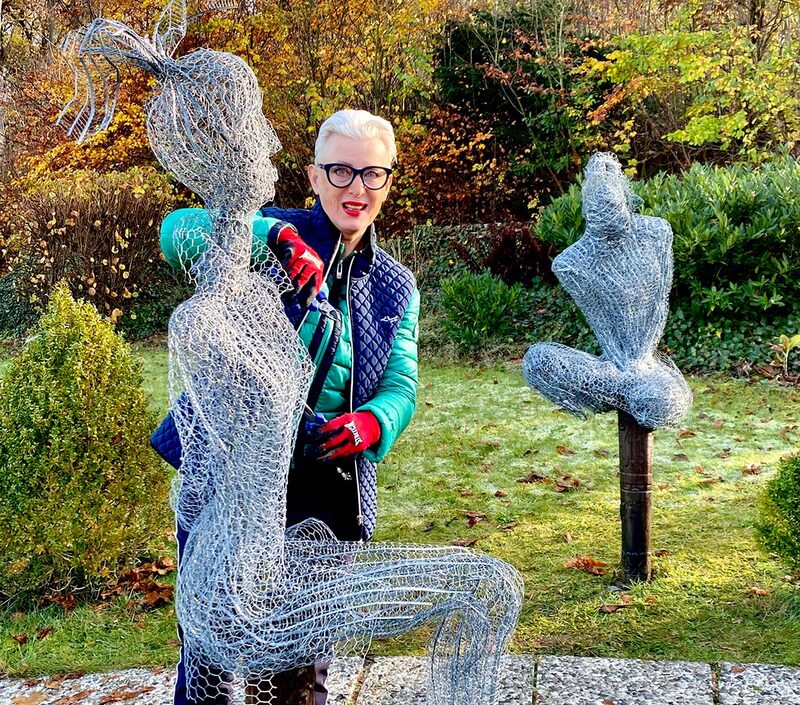 Die Künstlerin zwischen zwei lebensgroßen Drahtfiguren im Garten