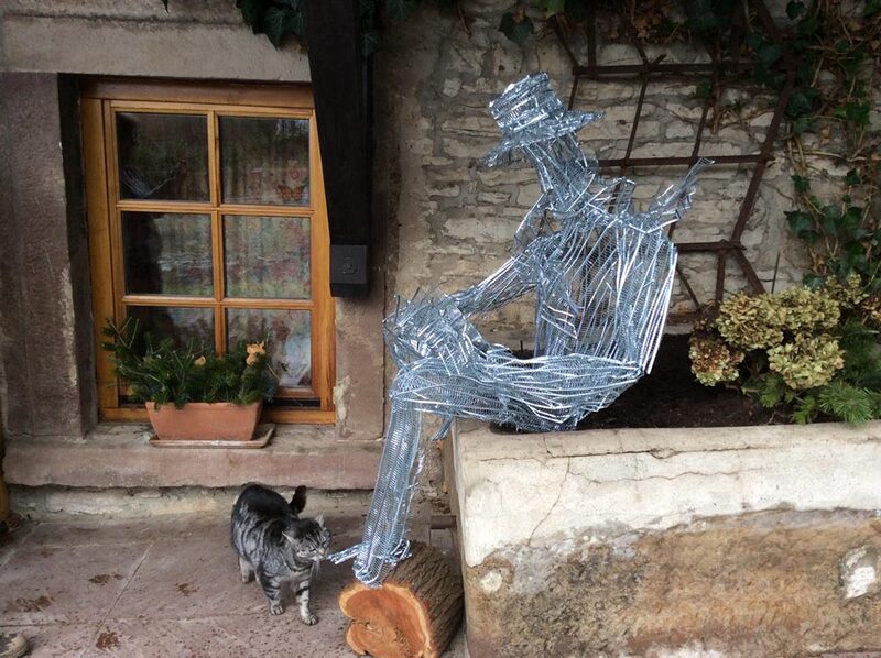 Sitzende Drahtfigur auf einem Baumstamm, davor zwei Katzen